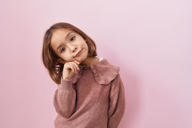 Menina hispânica em pé sobre fundo rosa, cara séria pensando em questão com a mão no queixo pensativa sobre ideia confusa