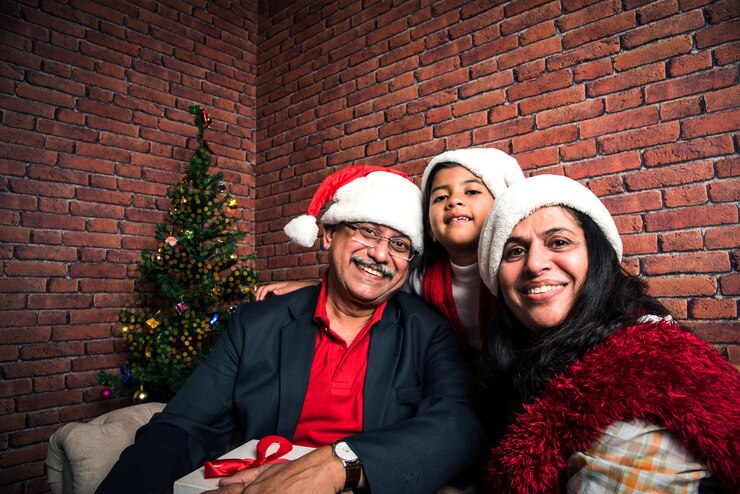 Foto menina indiana asiática fofa celebrando o natal com os avós e a árvore de natal, sentada no sofá, usando chapéu de papai noel e muitos presentes