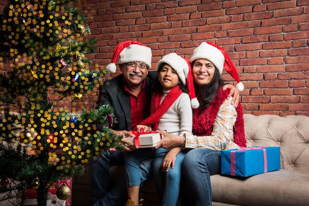 Menina indiana asiática fofa celebrando o Natal com os avós e a árvore de Natal, sentada no sofá, usando chapéu de Papai Noel e muitos presentes