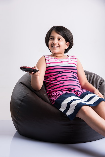 Menina indiana bonitinha assistindo televisão ou tv enquanto muda de canal usando o controle remoto, sentada sobre o saco de feijão, isolado sobre um fundo branco