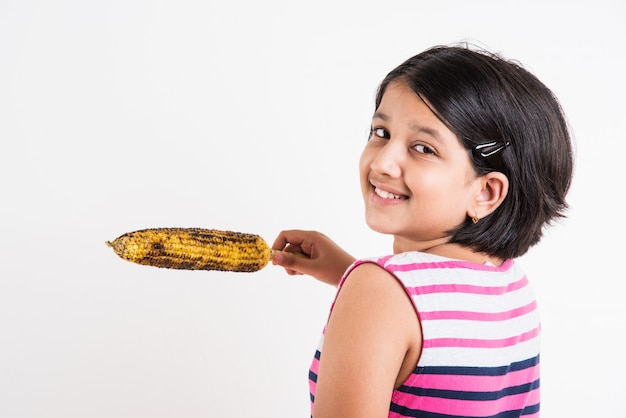Menina indiana bonitinha comendo milho grelhado ou bhutta, em pé, isolada sobre um fundo branco