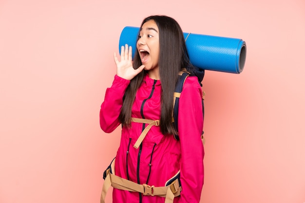Menina indiana jovem alpinista com uma mochila grande na parede rosa gritando com a boca aberta