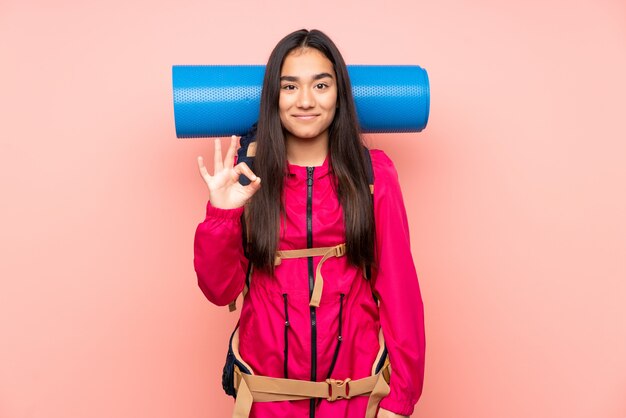 Menina indiana jovem alpinista com uma mochila grande na parede rosa, mostrando um sinal de ok com os dedos