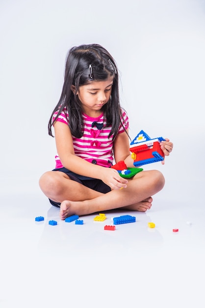 Menina indiana ou asiática bonitinha brincando com brinquedos ou blocos coloridos e se divertindo enquanto está sentada à mesa ou isolada sobre um fundo branco