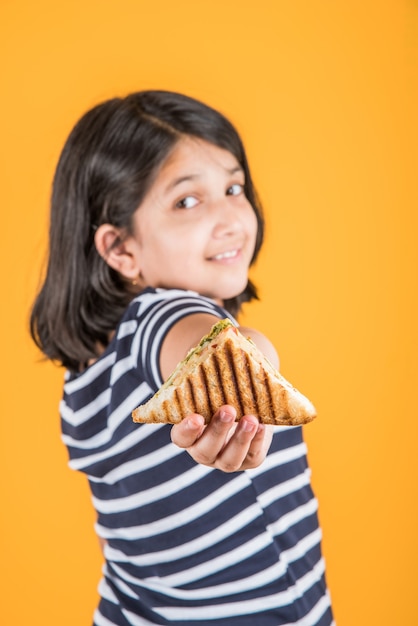 Menina indiana ou asiática bonitinha comendo hambúrguer saboroso, sanduíche ou pizza em um prato ou caixa. Permanente isolado sobre fundo azul ou amarelo.