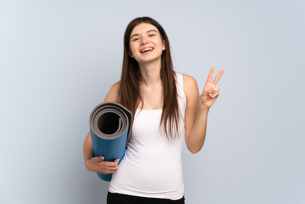 Menina indo para as aulas de ioga, segurando um tapete, sorrindo e mostrando o sinal da vitória