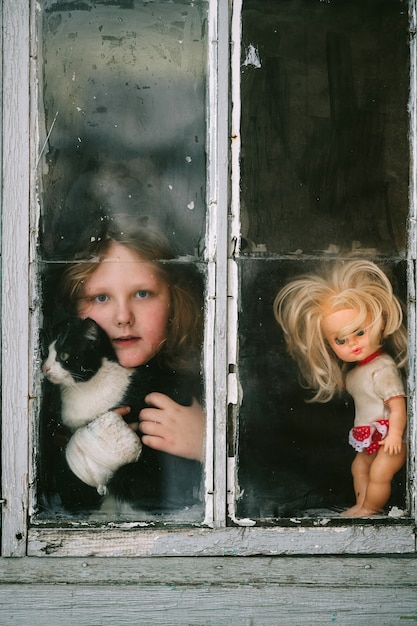 Menina isolada com seu gatinho e boneca olhando pela janela durante a pandemia de coronavírus