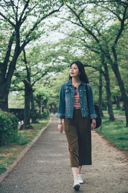 menina japonesa asiática de comprimento total caminha ao longo do caminho na floresta sob árvores verdes no parque primavera ao ar livre. turista de mulher aprecia o jardim da natureza na cidade, olhando de lado para relaxar. mochileiro feminino na passarela.