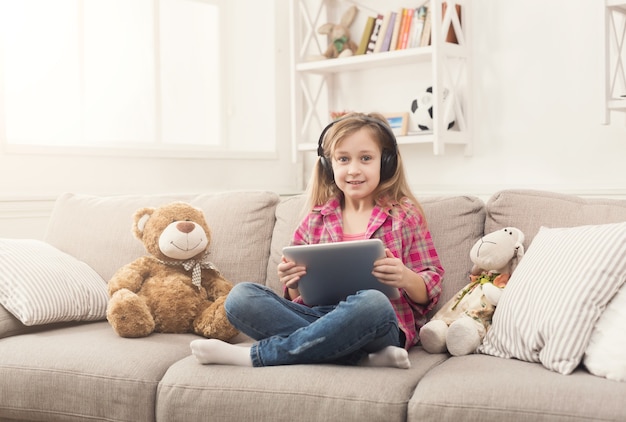 Menina jogando jogos online no tablet digital e ouvindo música em fones de ouvido. Criança do sexo feminino sentada no sofá com seus brinquedos. Rede social e conceito de educação online