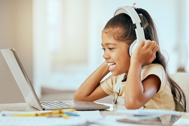 Menina laptop e fones de ouvido com sorriso música ou vídeo na mesa na aprendizagem e educação escolar em casa Computador infantil e feliz por podcast na internet web ou aplicativo por mesa em casa para aula online