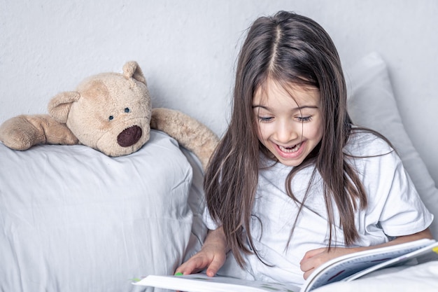 Menina lê um livro com um ursinho de pelúcia na cama de manhã