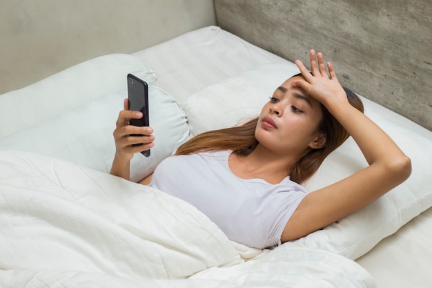 Menina linda asiática infeliz se sentindo triste usando o smartphone na cama