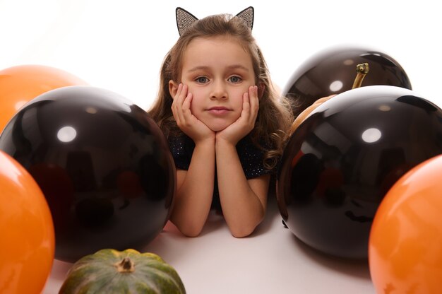 Menina linda criança com aro com orelhas de gato olha para a câmera deitada ao lado de balões pretos e laranja coloridos em um fundo branco com espaço de cópia. Conceito de halloween