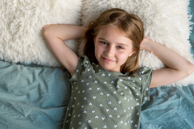 Menina linda criança com cabelo loiro deitada na cama e olhando para a câmera no quarto com ...