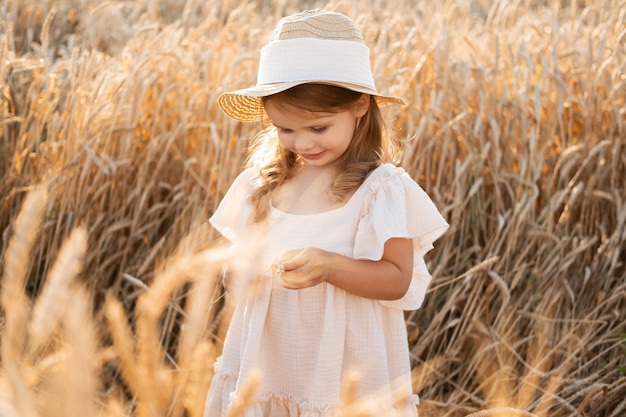 Menina linda criança com chapéu de palha e vestido bege de musselina no campo de trigo no pôr do sol.