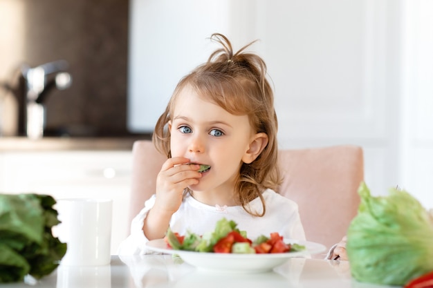 Menina linda criança comendo salada de vegetais crus frescos na cozinha branca