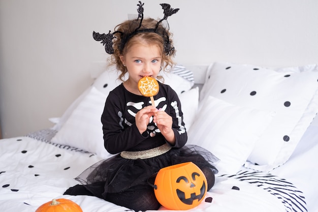 Menina linda criança fantasiada de bruxa comendo biscoitos decorados para o quarto de halloween.