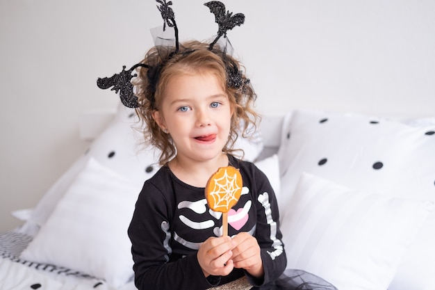 Menina linda criança fantasiada de bruxa comendo biscoitos decorados para o quarto de halloween.