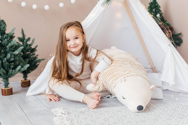 Menina linda de pijama branco brinca com um ursinho de pelúcia perto do quarto das crianças da tenda com c