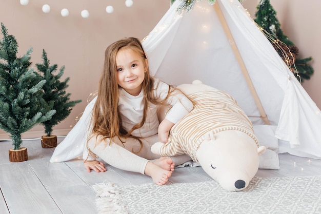 Menina linda de pijama branco brinca com um ursinho de pelúcia perto do teepee children39s room com decorações e decorações de árvores de natal