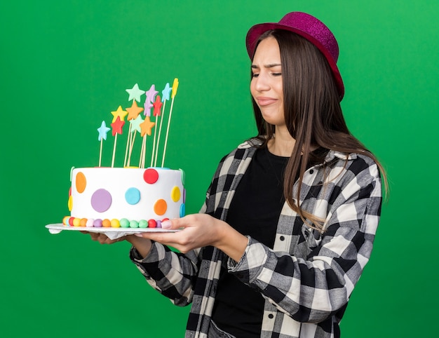 Menina linda e desagradável com chapéu de festa segurando e olhando para um bolo