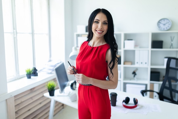 Foto menina linda em um terno vermelho está de pé no escritório e está segurando um lápis na mão