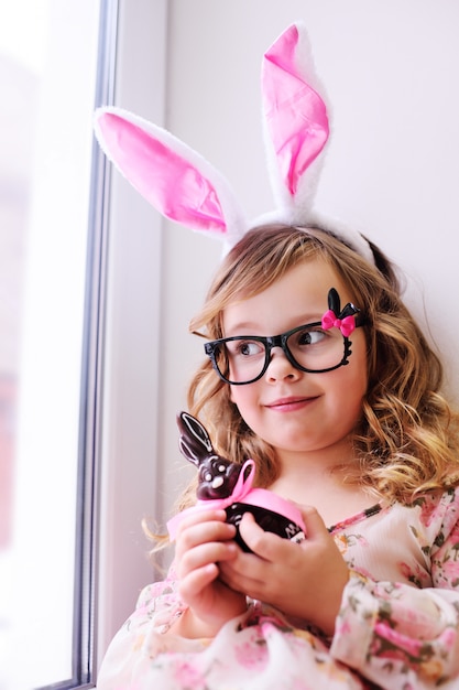 Menina linda em um vestido inteligente com orelhas de coelho na cabeça, segurando um coelhinho da Páscoa de chocolate