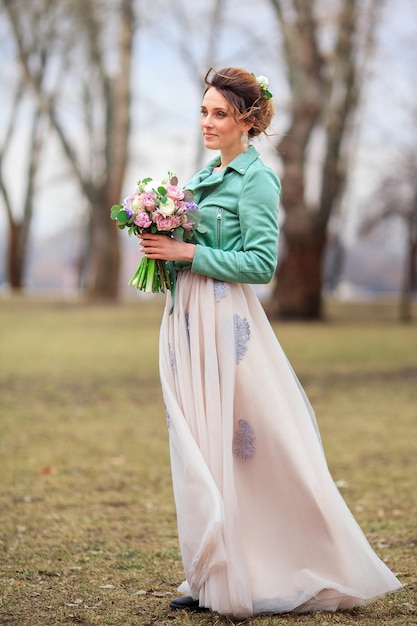 Menina linda em um vestido longo e uma jaqueta verde e com um buquê de flores nas mãos dela caminha entre as árvores do parque. ela está feliz e sorrindo.