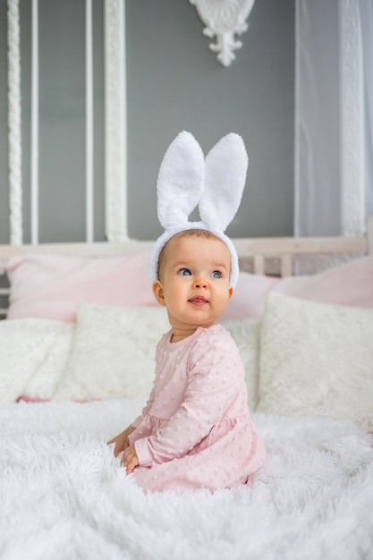 Menina linda em um vestido rosa com uma faixa na cabeça e orelhas de coelho está sentada na cama no quarto