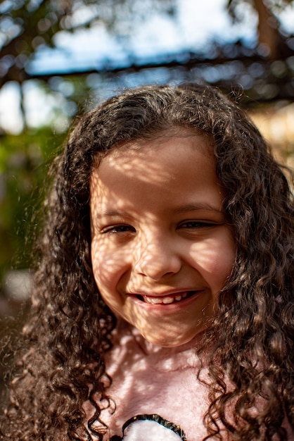 Menina, linda garotinha com cabelo encaracolado sob a sombra de árvores feliz, pequena profundidade de campo, foco seletivo.