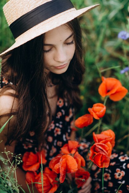 Menina linda jovem morena modelo com um buquê de papoilas vermelhas em um chapéu fechou os olhos e aprecia o aroma das flores. Retrato feminino no verão.