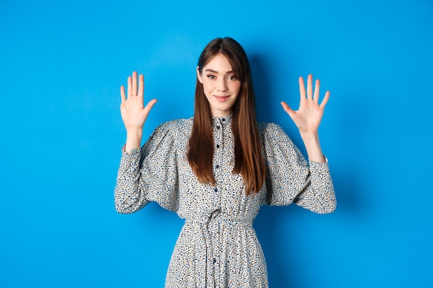 Menina linda num vestido com cabelo comprido natural, mostrando o número dez com os dedos e sorrindo, em pé contra o azul.