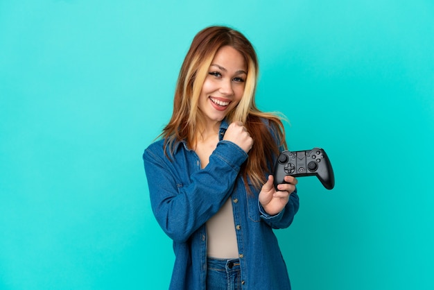 Menina loira adolescente brincando com um controle de videogame sobre uma parede isolada comemorando uma vitória