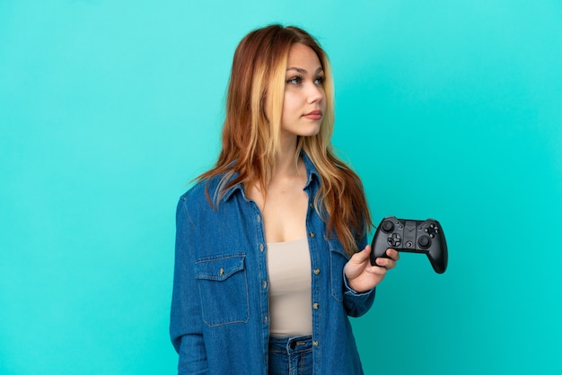 Menina loira adolescente brincando com um controle de videogame sobre uma parede isolada olhando para o lado