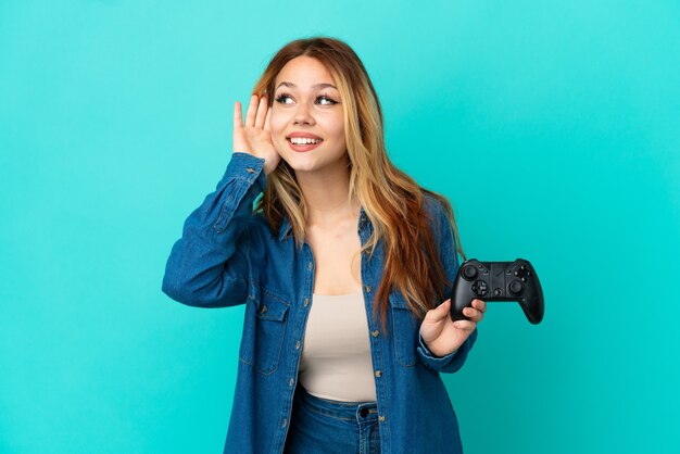 Menina loira adolescente brincando com um controle de videogame sobre uma parede isolada, ouvindo algo colocando a mão na orelha