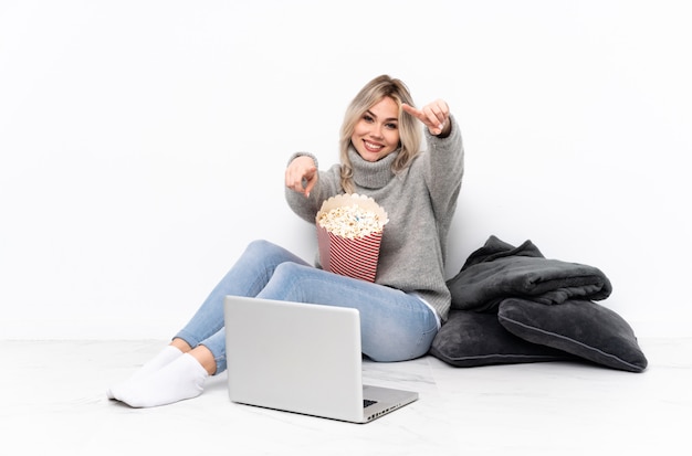 Menina loira adolescente comendo pipoca enquanto assiste a um filme no laptop aponta o dedo para você enquanto sorrindo