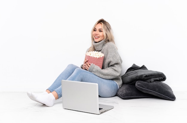 Menina loira adolescente comendo pipoca enquanto assiste a um filme no laptop feliz e sorridente