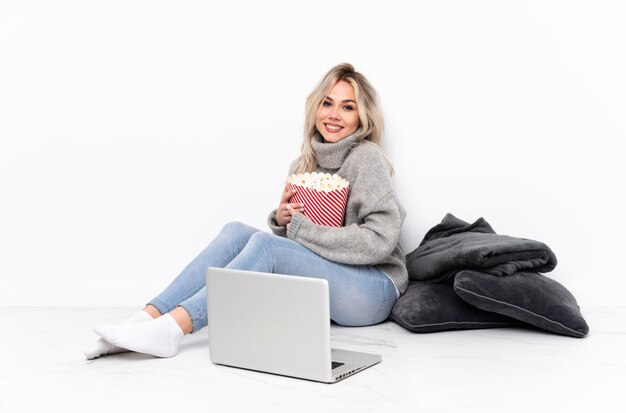 Menina loira adolescente comendo pipoca enquanto assiste a um filme no laptop, mantendo os braços cruzados na posição frontal