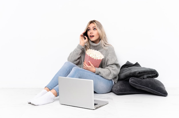 Menina loira adolescente comendo pipoca enquanto assiste a um filme no laptop segurando café para levar embora e um celular