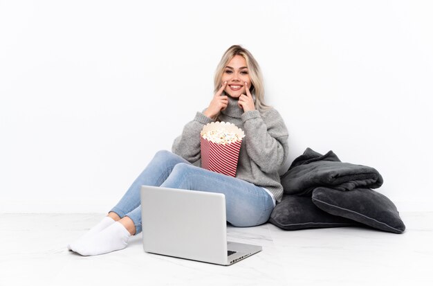 Menina loira adolescente comendo pipoca enquanto assiste a um filme no laptop sorrindo com uma expressão feliz e agradável