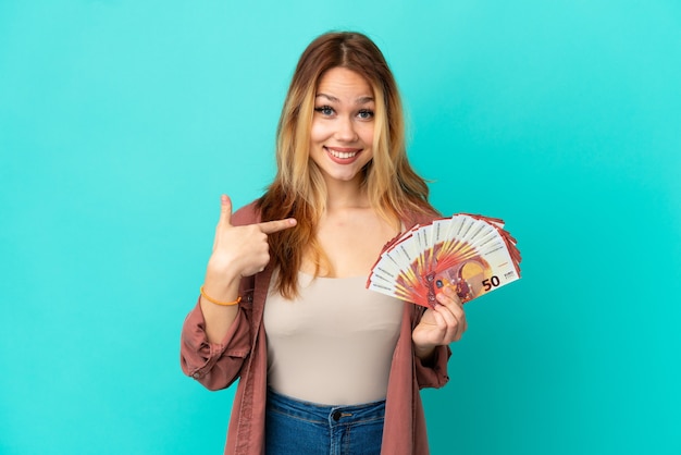 Menina loira adolescente pegando muitos euros sobre um fundo azul isolado com expressão facial de surpresa