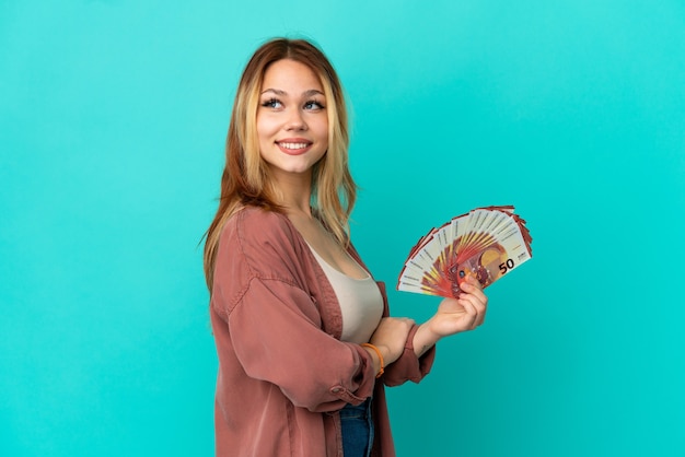 Menina loira adolescente pegando muitos euros sobre um fundo azul isolado com os braços cruzados e feliz