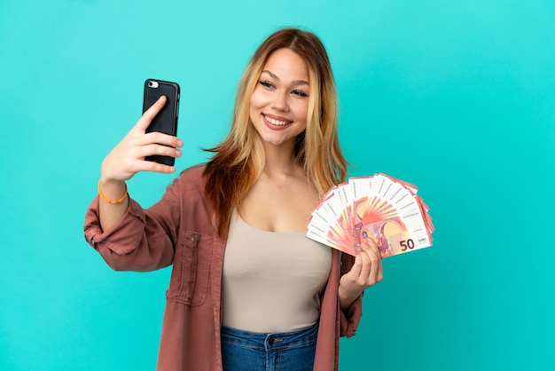 Menina loira adolescente pegando muitos euros sobre um fundo azul isolado fazendo uma selfie