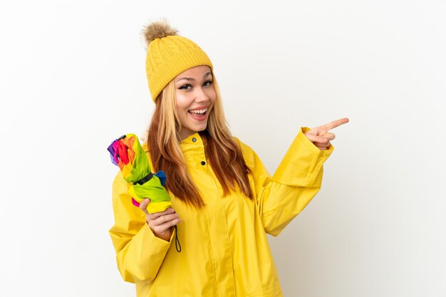 Menina loira adolescente vestindo um casaco à prova de chuva sobre um fundo branco isolado apontando o dedo para o lado e apresentando um produto