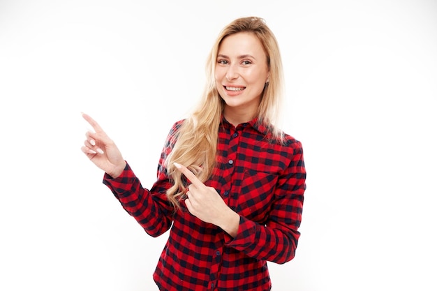 Menina loira alegre em uma camisa vermelha xadrez sorrindo aponta o dedo para o espaço vazio da cópia para texto ou produto isolado no banner de publicidade de fundo branco
