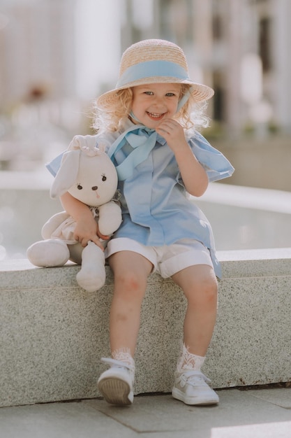 menina loira bonitinha com um chapéu de palha e um vestido azul brinca com um coelho de pelúcia ao ar livre