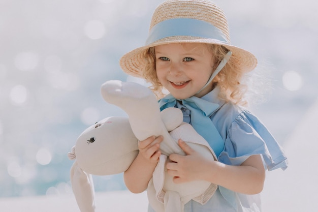 menina loira bonitinha de vestido azul e chapéu de palha jogando perto de fonte com coelhinho de pelúcia, cartão