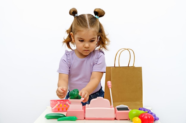 Menina loira brinca na loja pesa legumes em balanças de brinquedos coleta pedido em pacote de artesanato caixa registradora infantil jogos caseiros para crianças comida online pedidos entrega de alimentos