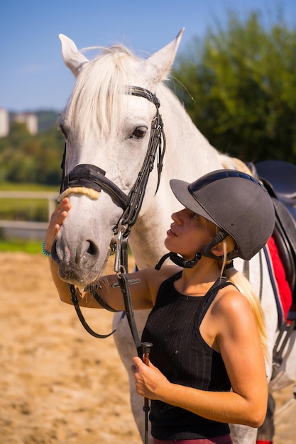 Menina loira caucasiana acariciando um cavalo branco em um passeio a cavalo, vestida de cavaleiro preto com chapéu de segurança