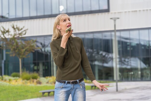 Menina loira caucasiana empreendedora em um parque empresarial, fazendo uma anotação de áudio com o telefone, um moderno local de trabalho entre a natureza